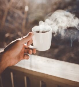 Photo shows  hand holding a cup of steaming black coffee over a wooden railing looking off into an unfocused background with leafless trees, like winter without snow 